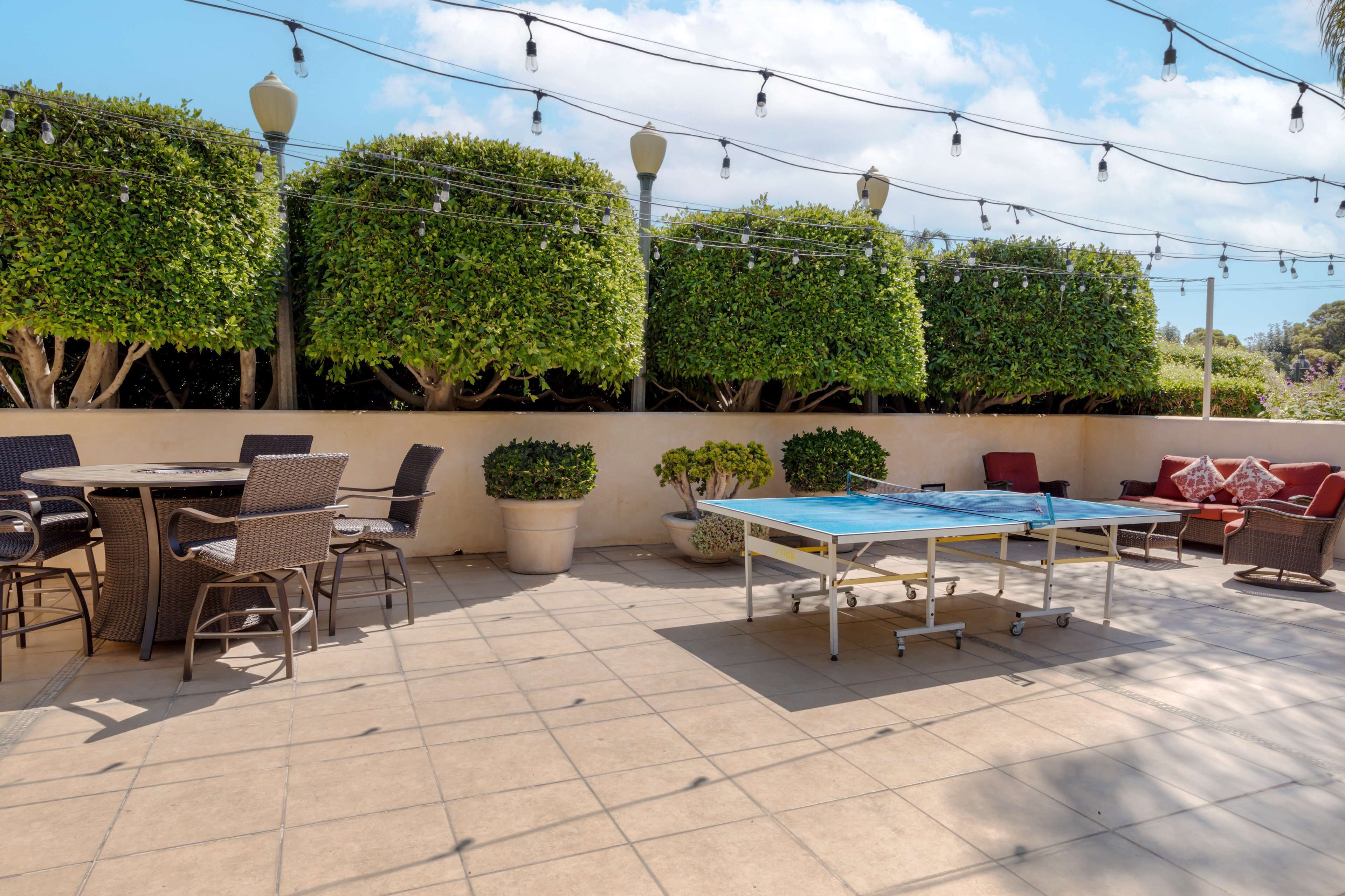 An outdoor patio area with a round table and chairs, a ping pong table, potted plants, and string lights.