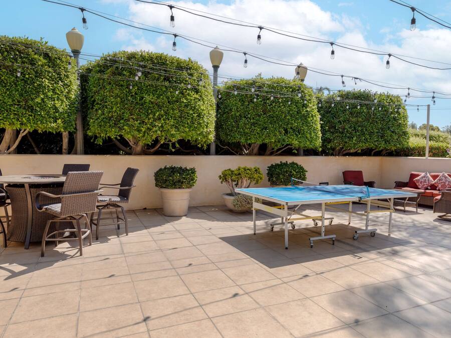 An outdoor patio area with a round table and chairs, a ping pong table, potted plants, and string lights.