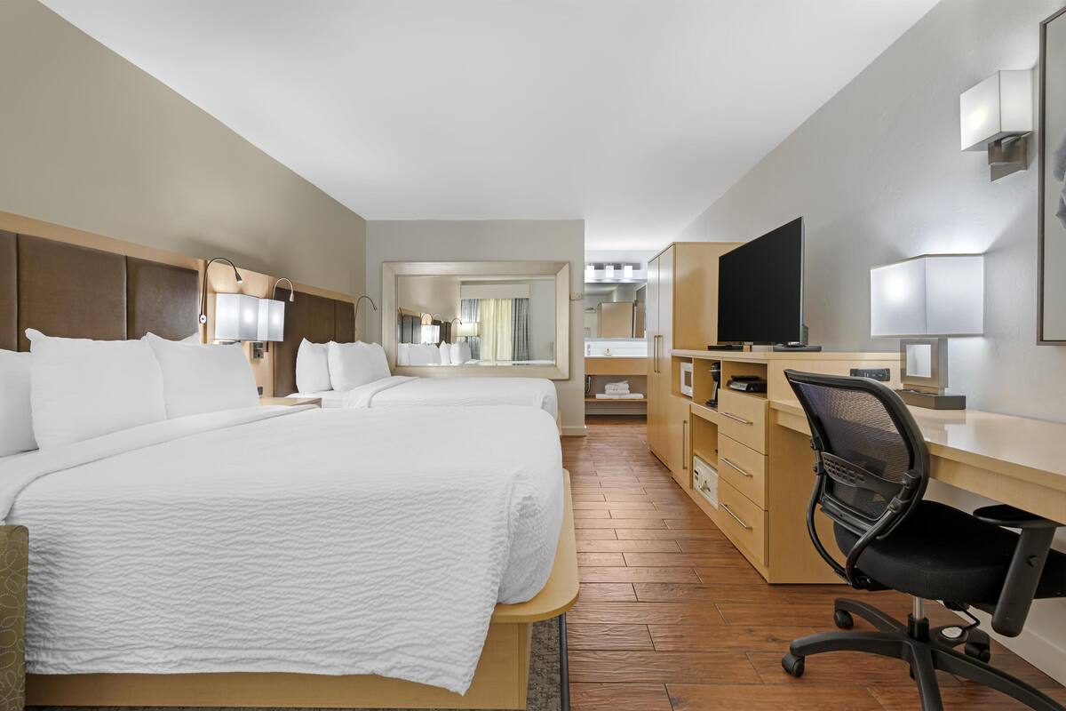 A hotel room with two beds, a desk, a chair, a TV, and a bathroom visible through a door.