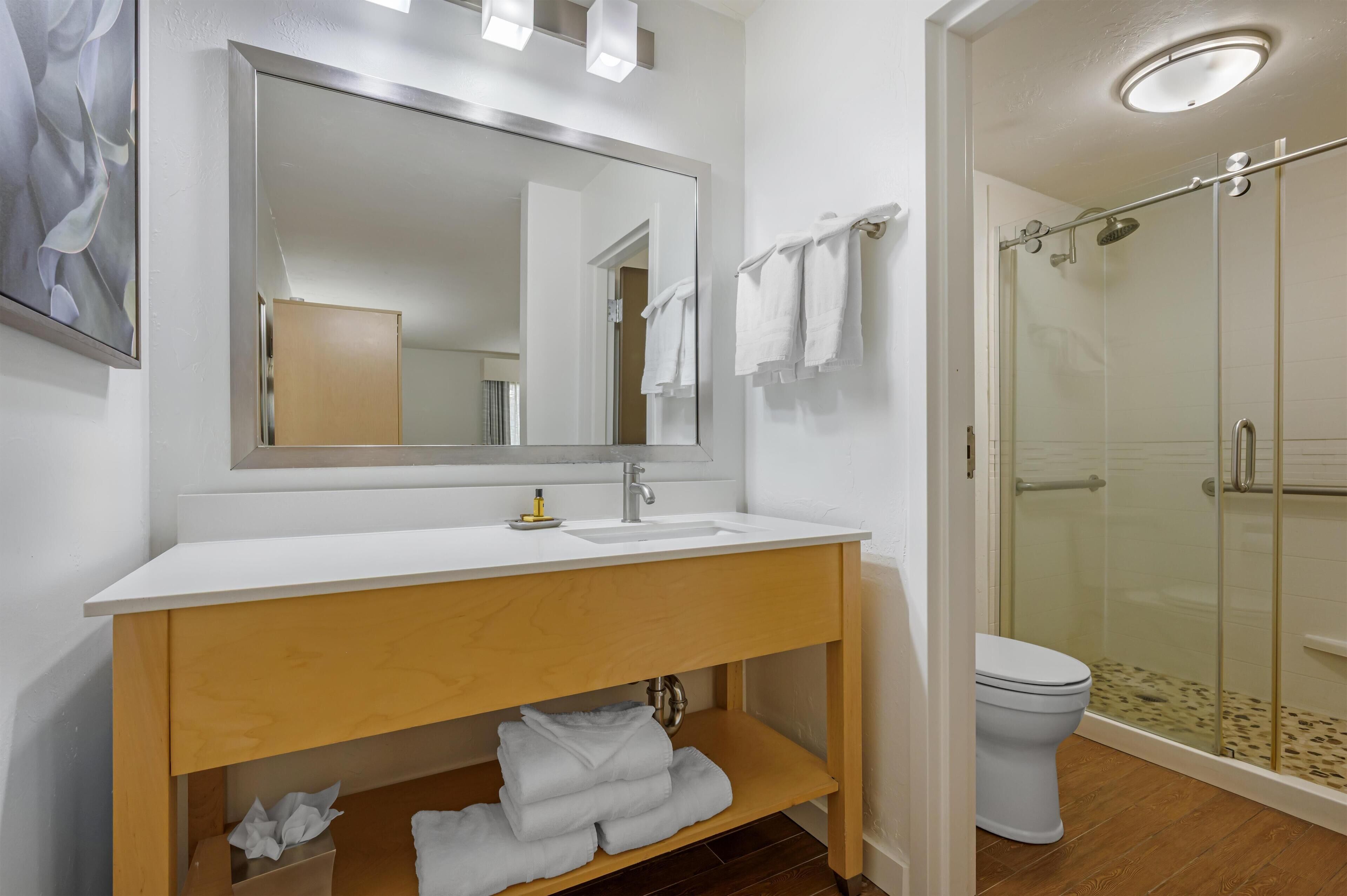 A modern bathroom with a large mirror, white sink, wooden vanity, white towels, and a glass-enclosed shower.