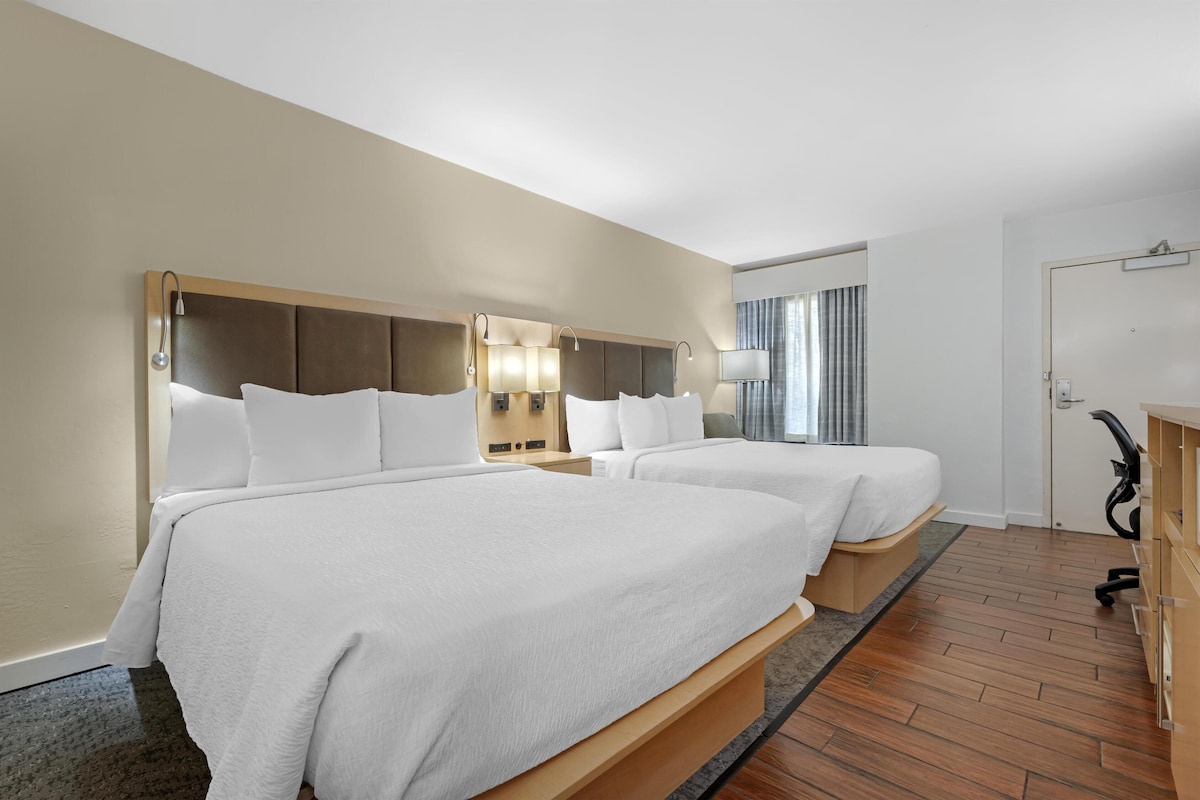 A hotel room with two beds, a wooden headboard, bedside lamps, a desk, and a chair.