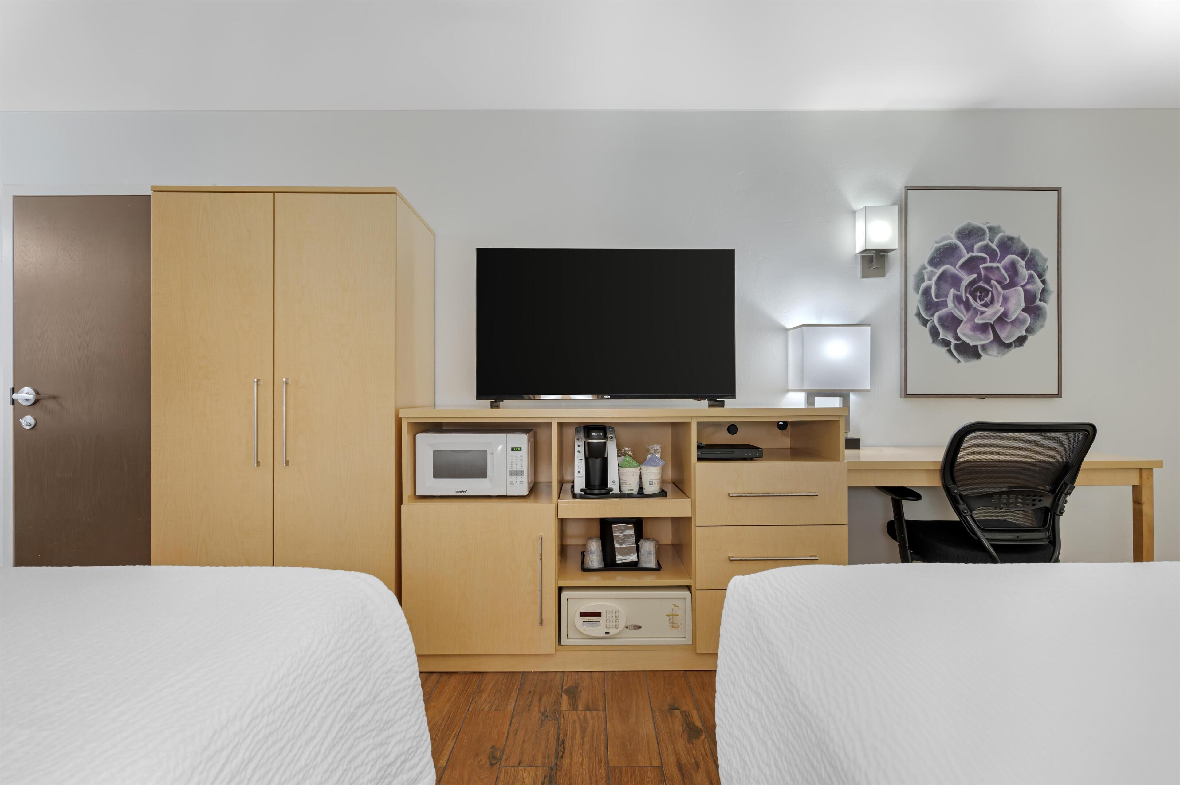 A hotel room with two beds, a desk with a flat-screen TV, a microwave, a coffee maker, and a chair.