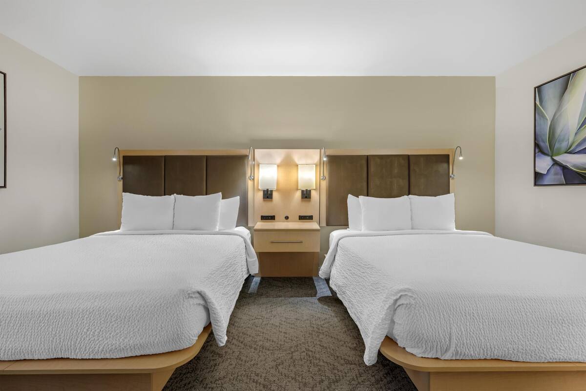 A hotel room with two single beds, a wooden headboard, a nightstand with a lamp, and a wall-mounted mirror.