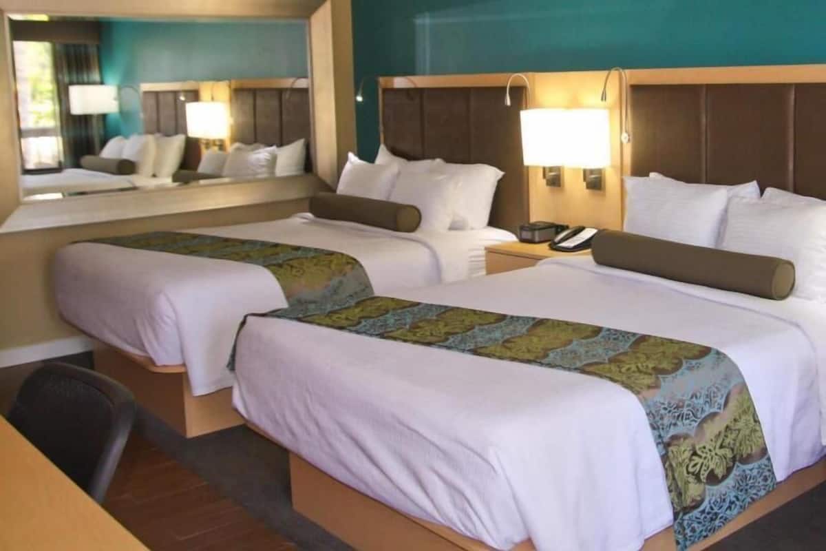 A hotel room with two beds, a desk, a chair, a mirror, and a nightstand with a lamp.