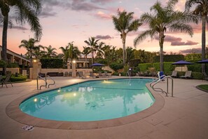 A swimming pool with lounge chairs and umbrellas, surrounded by palm trees and a building.