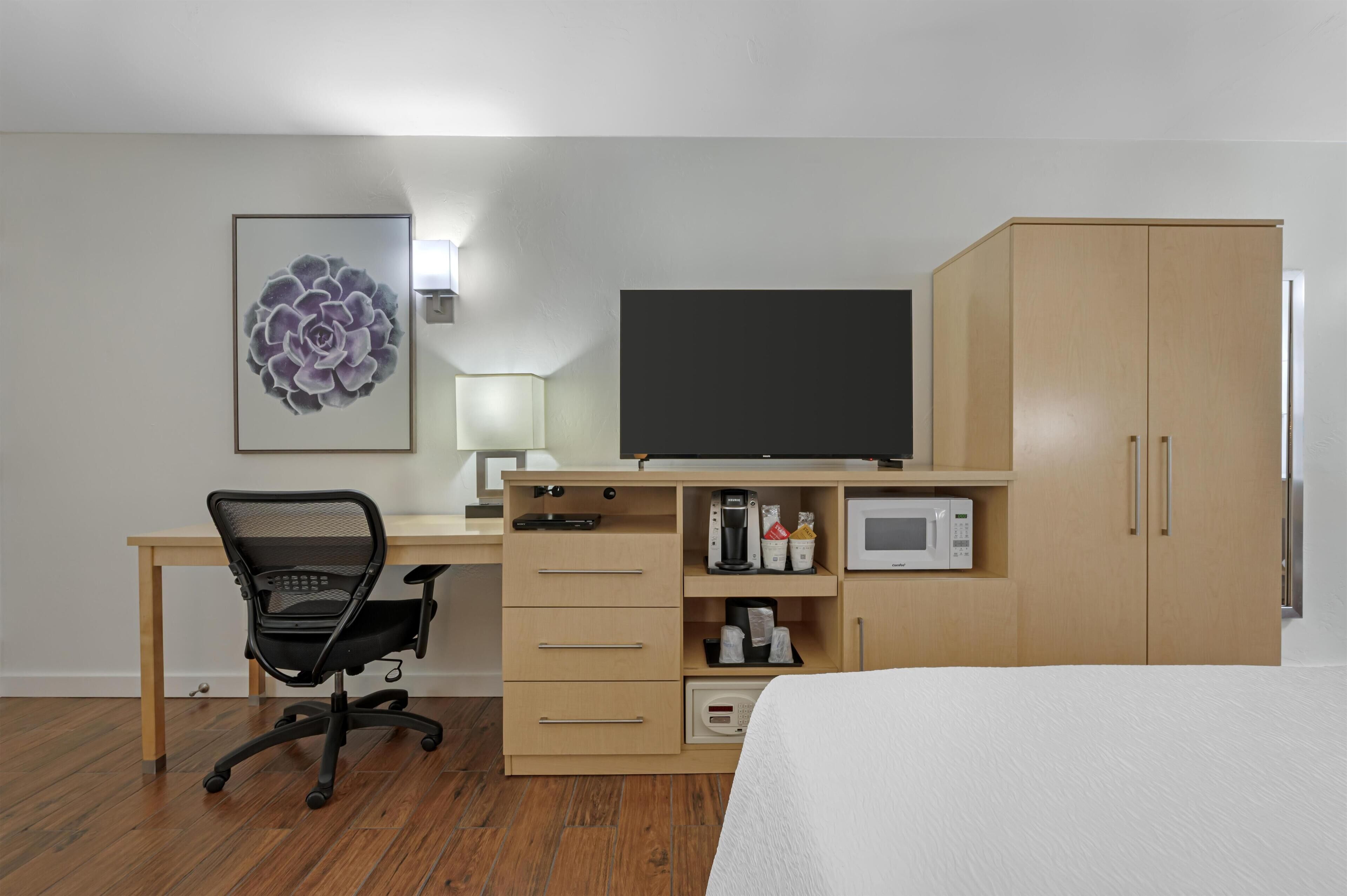 A hotel room with a wooden desk, a black office chair, a flat-screen TV, a microwave, a coffee maker, and a wardrobe.