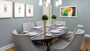 A dining room with a round table set for six, surrounded by six grey upholstered chairs with beaded trim. There are three framed artworks on the wall, a chandelier above the table, and a potted plant on the table.