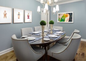 Comedor con una mesa redonda dispuesta para seis, rodeada por seis sillas grises tapizadas con adornos perlados. Hay tres obras de arte enmarcadas en la pared, un candelabro sobre la mesa y una planta en maceta sobre la misma.