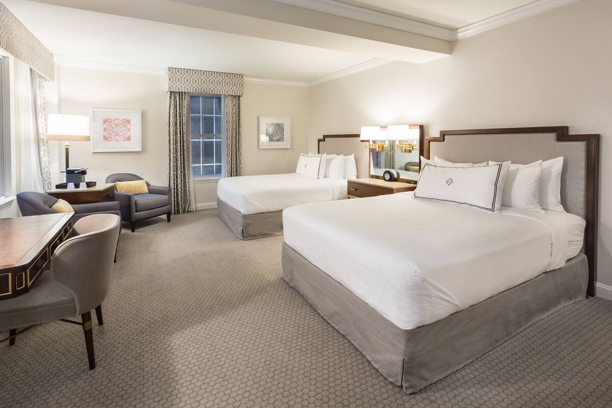 A hotel room with two beds, a desk, a chair, a dresser, and a large window with curtains.