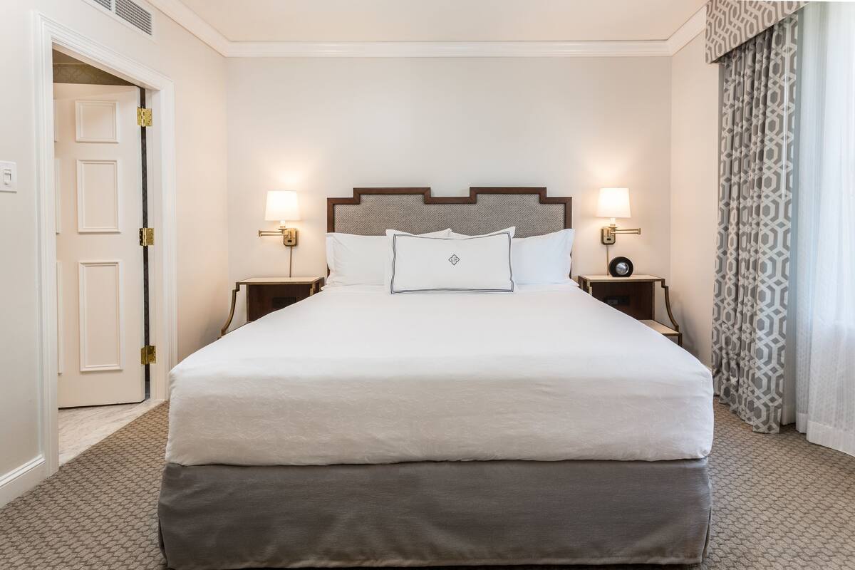 A hotel room with a large bed, two bedside tables with lamps, a headboard with a geometric pattern, and a patterned curtain.