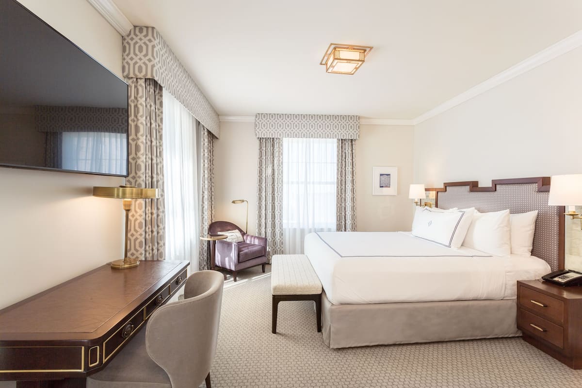 A hotel room with a large bed, a desk, a chair, a bedside table, and a TV.
