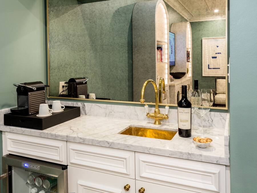 Un baño con lavamanos de mármol, grifo dorado y una botella de vino. Hay armarios blancos y un espejo sobre el lavamanos.