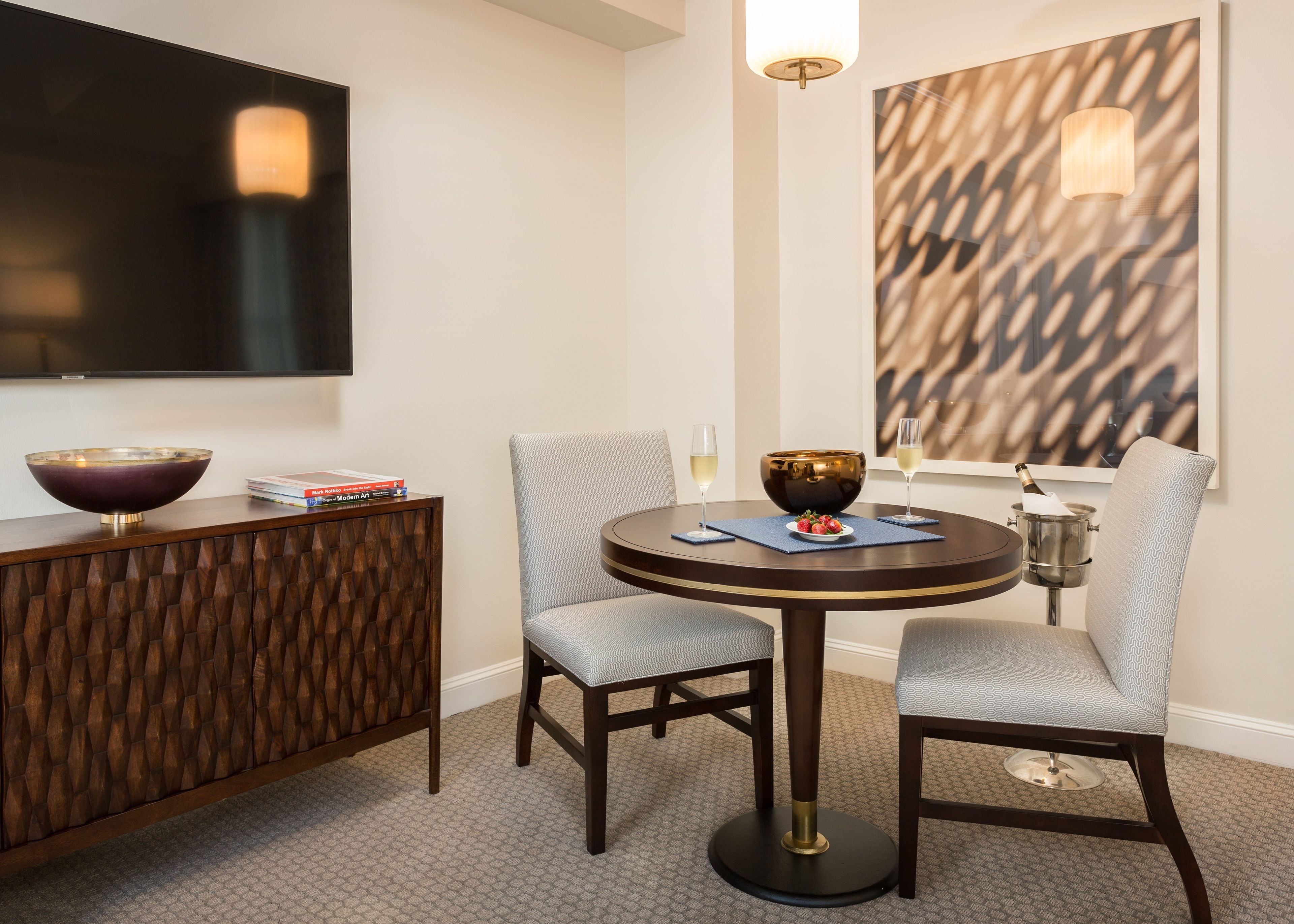 A modern dining area with a round table, two chairs, a bowl, a bottle of champagne, and a book on a side table.