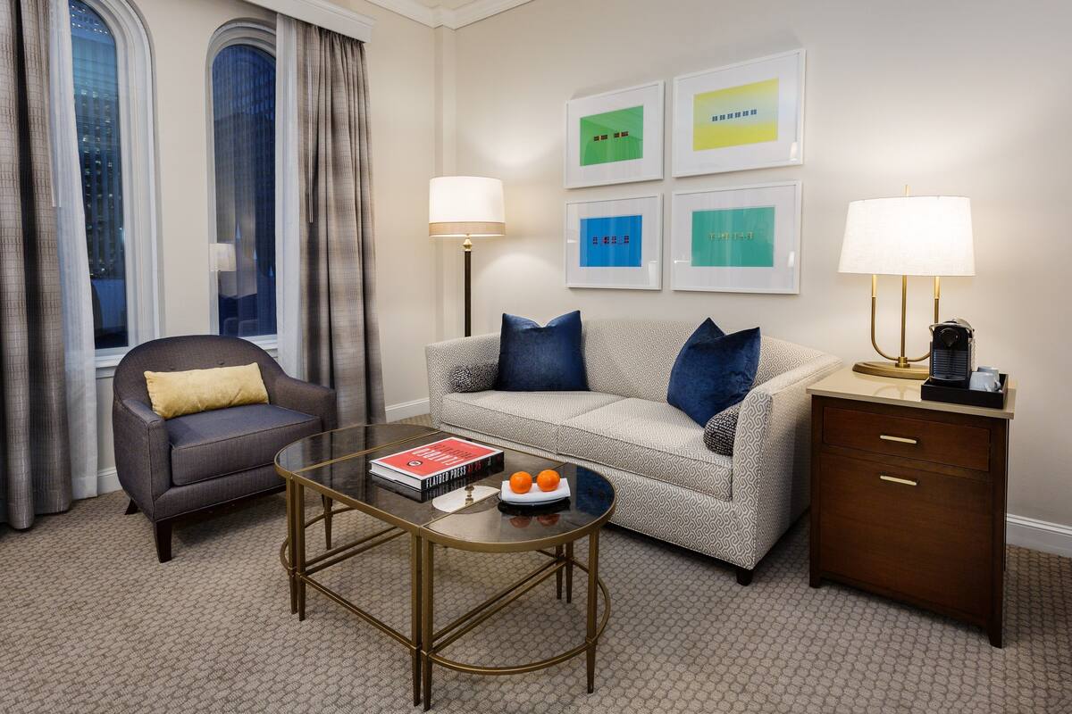A modern hotel room with a grey sofa, a small round table with a book and oranges, a side table with a lamp, and a nightstand with drawers.