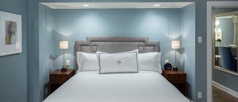A large bed with a grey headboard, two bedside tables with lamps, and a framed picture on the wall.