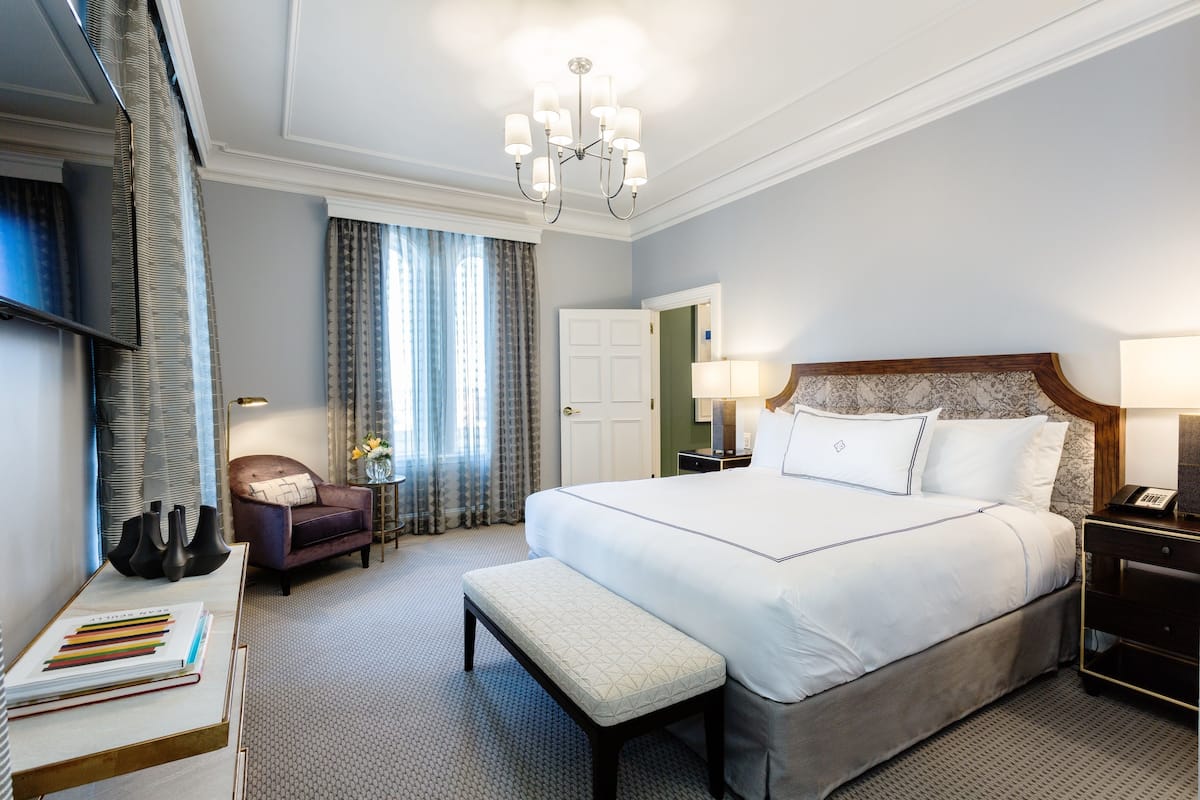 A hotel room with a large bed, a bench, a desk with books, a chair, a TV, and a chandelier.