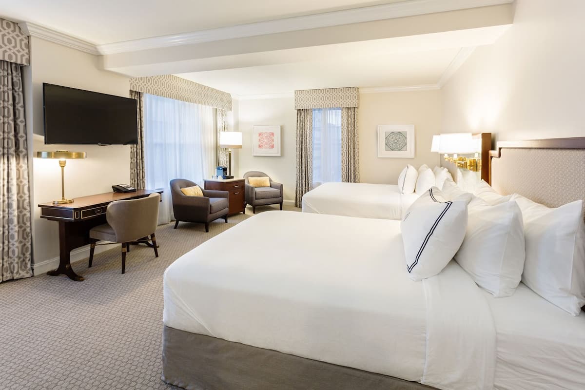 A hotel room with a large bed, a desk with a lamp, two armchairs, a TV, and a minibar.