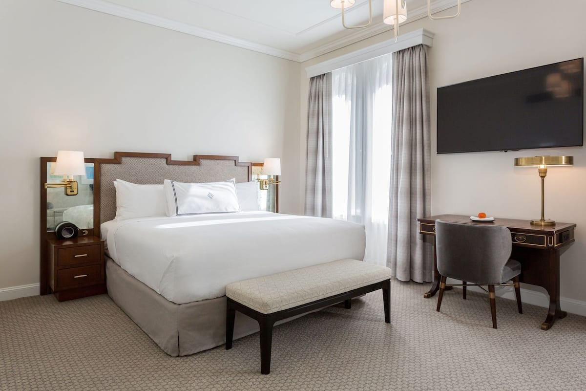 A hotel room with a large bed, bedside tables, a desk with a chair, a television, and a window with curtains.