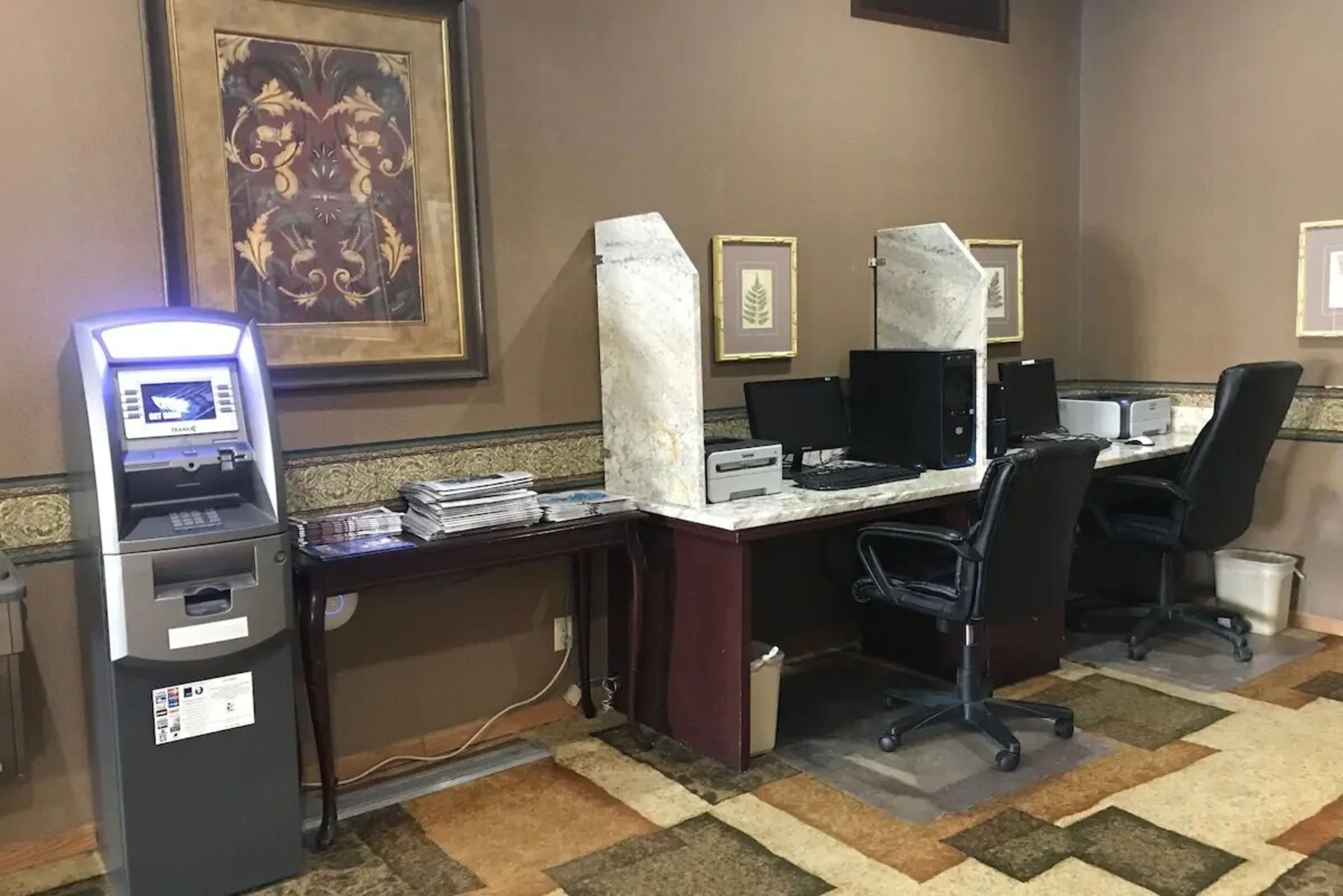 Une chambre d’hôtel équipée d’un bureau, d’un ordinateur et d’un distributeur automatique de billets. Les murs sont ornés de tableaux encadrés et le sol est recouvert d’une moquette à motifs.