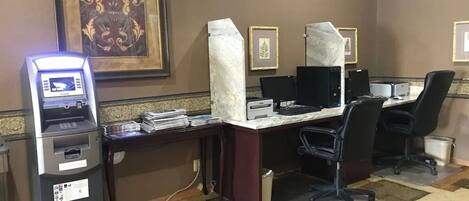 Une chambre d’hôtel équipée d’un bureau, d’un ordinateur et d’un distributeur automatique de billets. Les murs sont ornés de tableaux encadrés et le sol est recouvert d’une moquette à motifs.