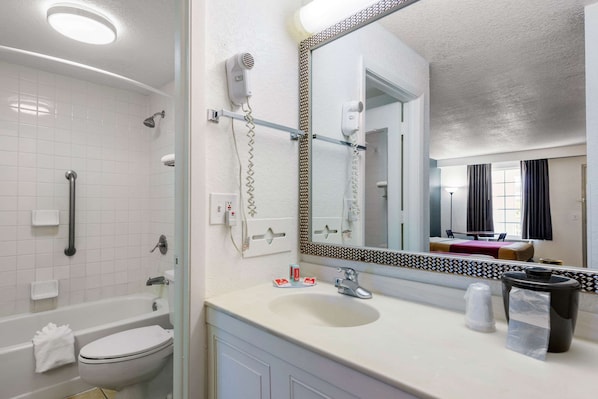 Une salle de bain moderne équipée d’une baignoire, de toilettes et d’un lavabo. On y trouve un sèche-cheveux fixé au mur, un miroir avec un cadre décoratif et une poubelle sur le comptoir du lavabo.