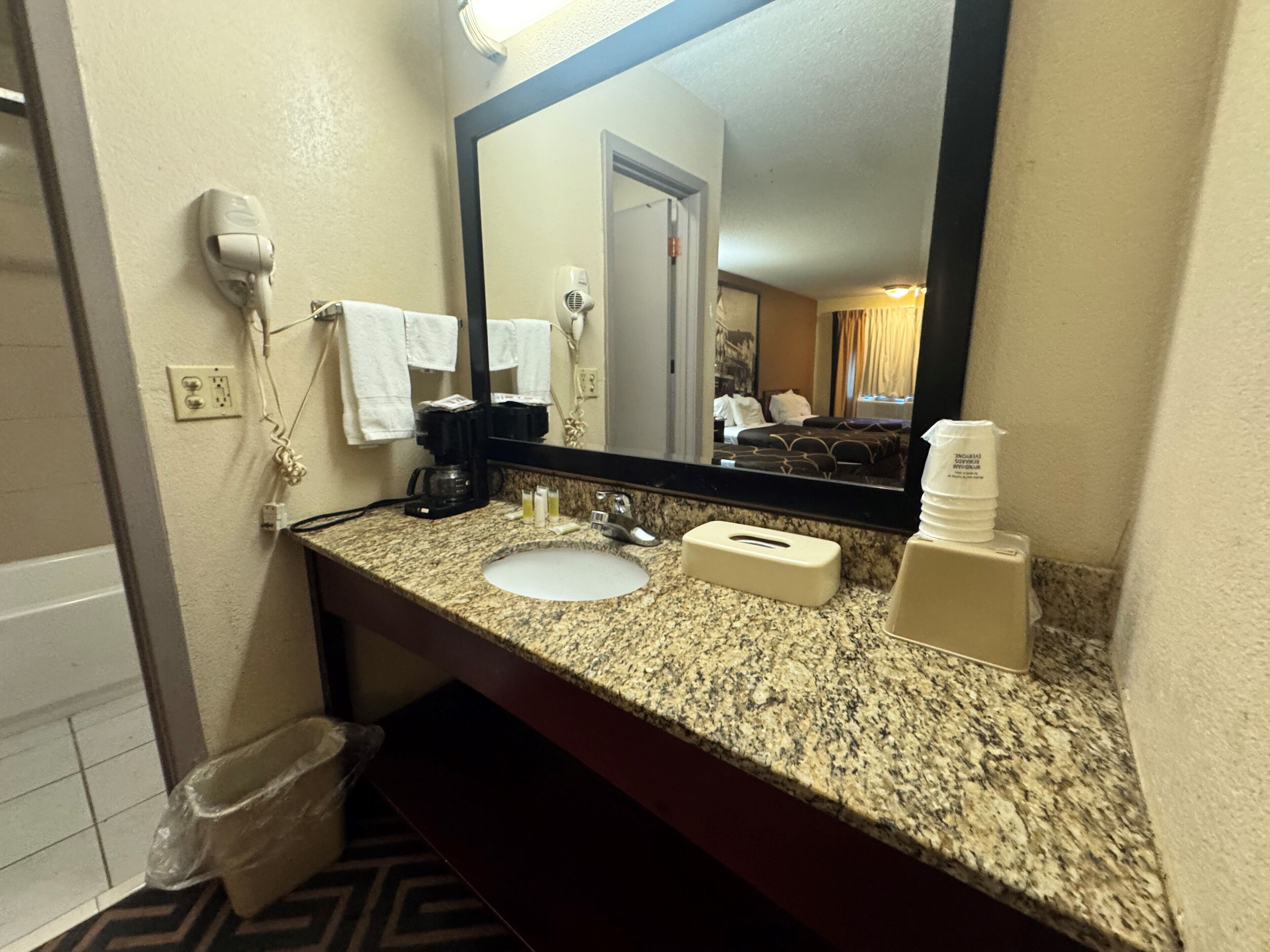 A hotel bathroom with a granite countertop, a sink, a mirror, a hair dryer, a towel rack, and a trash bin.