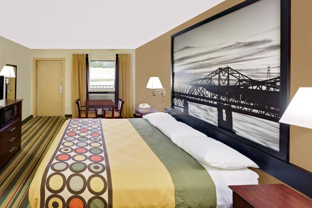Habitación de hotel con cama, escritorio, silla, lámpara y una ventana grande con cortinas.