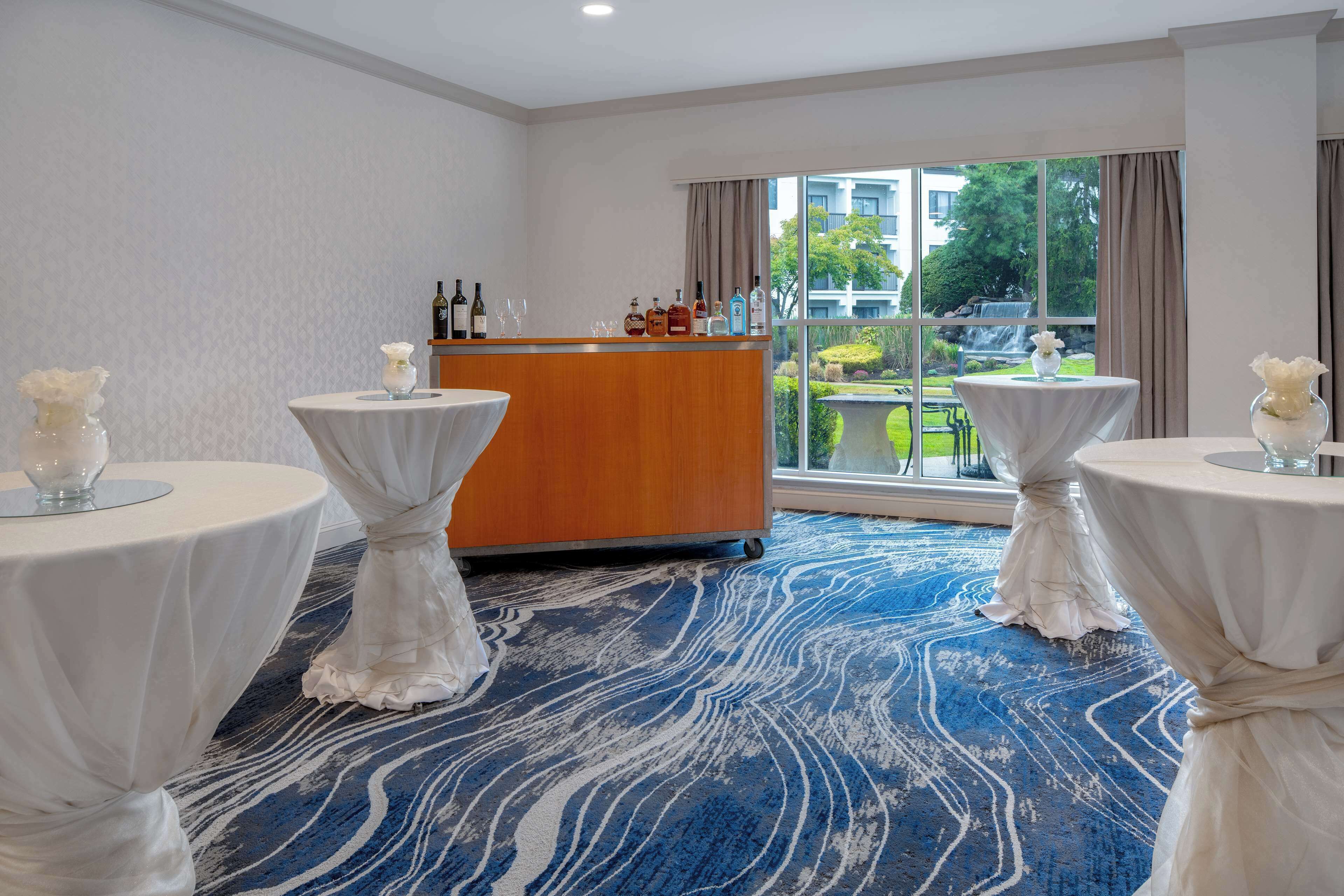 Una habitación de hotel con un ventanal grande, un mostrador de recepción de madera y tres mesas redondas con manteles blancos.