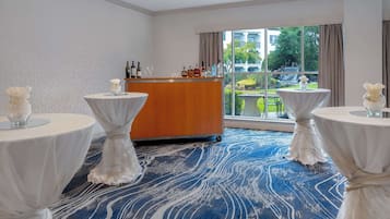 Una habitación de hotel con un ventanal grande, un mostrador de recepción de madera y tres mesas redondas con manteles blancos.