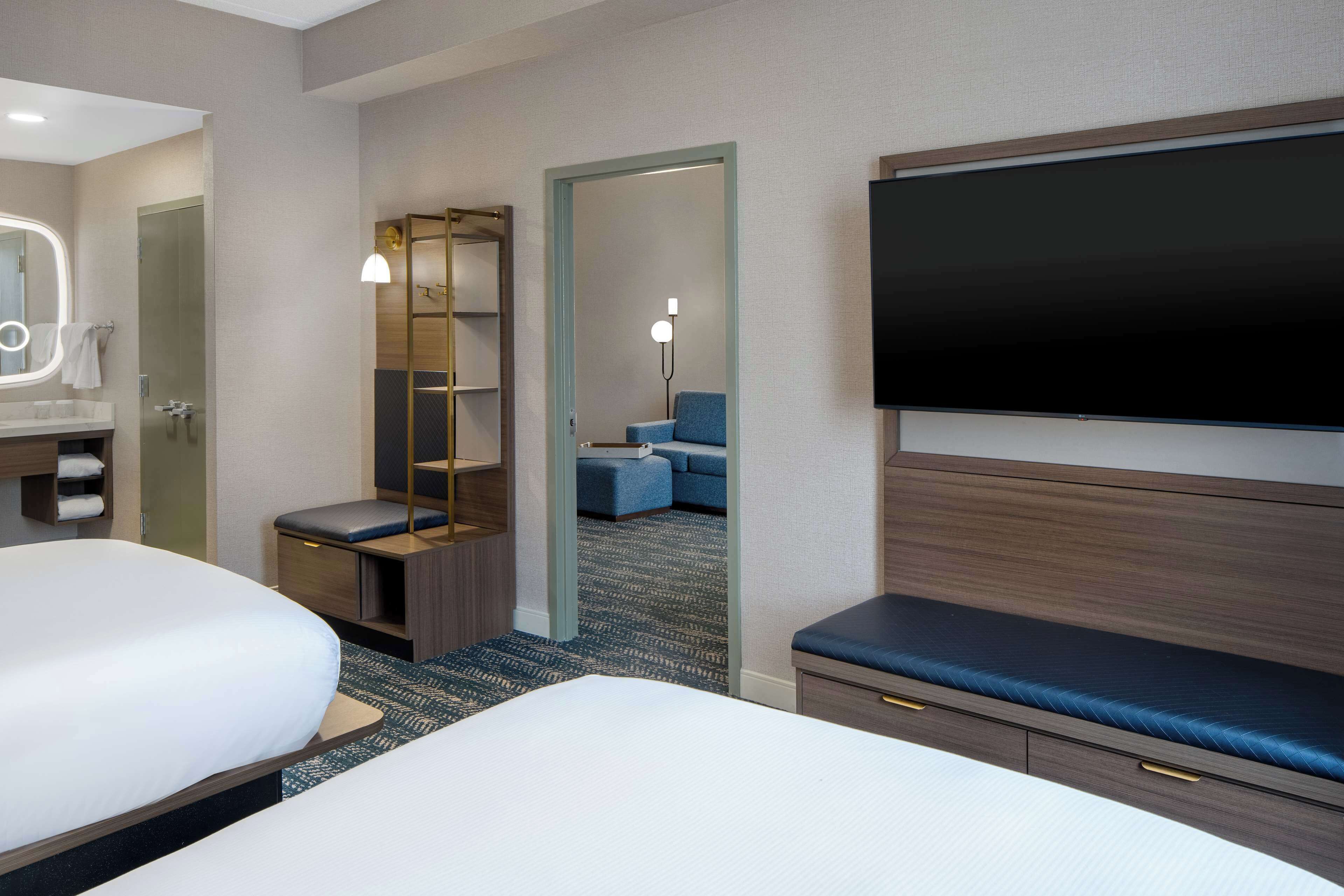 Quarto de hotel com duas camas, televisão, escrivaninha, espelho e banheiro.