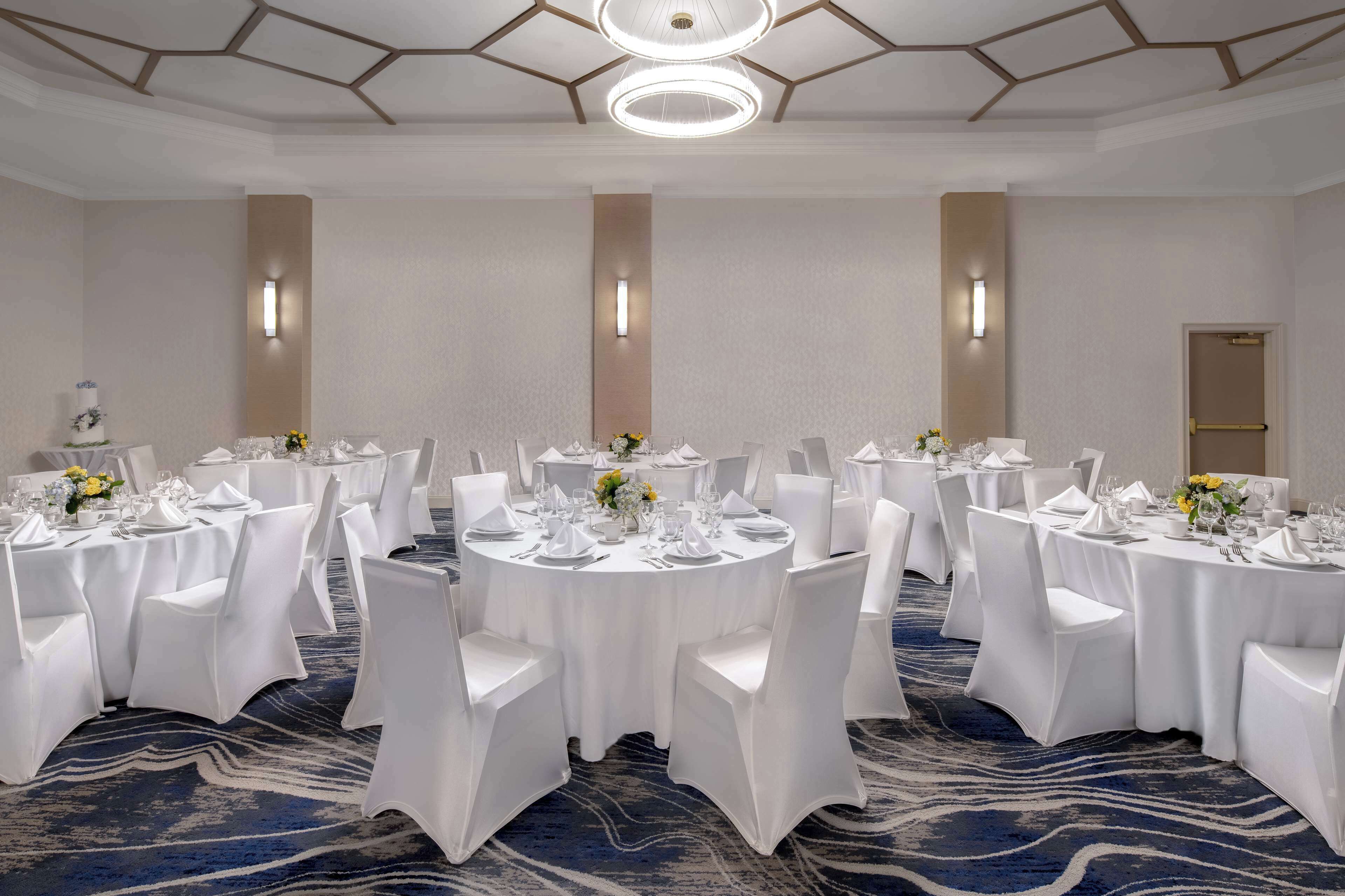 Un salón de conferencias con varias mesas redondas, cada una cubierta con mantel blanco, sillas y centros de mesa florales.