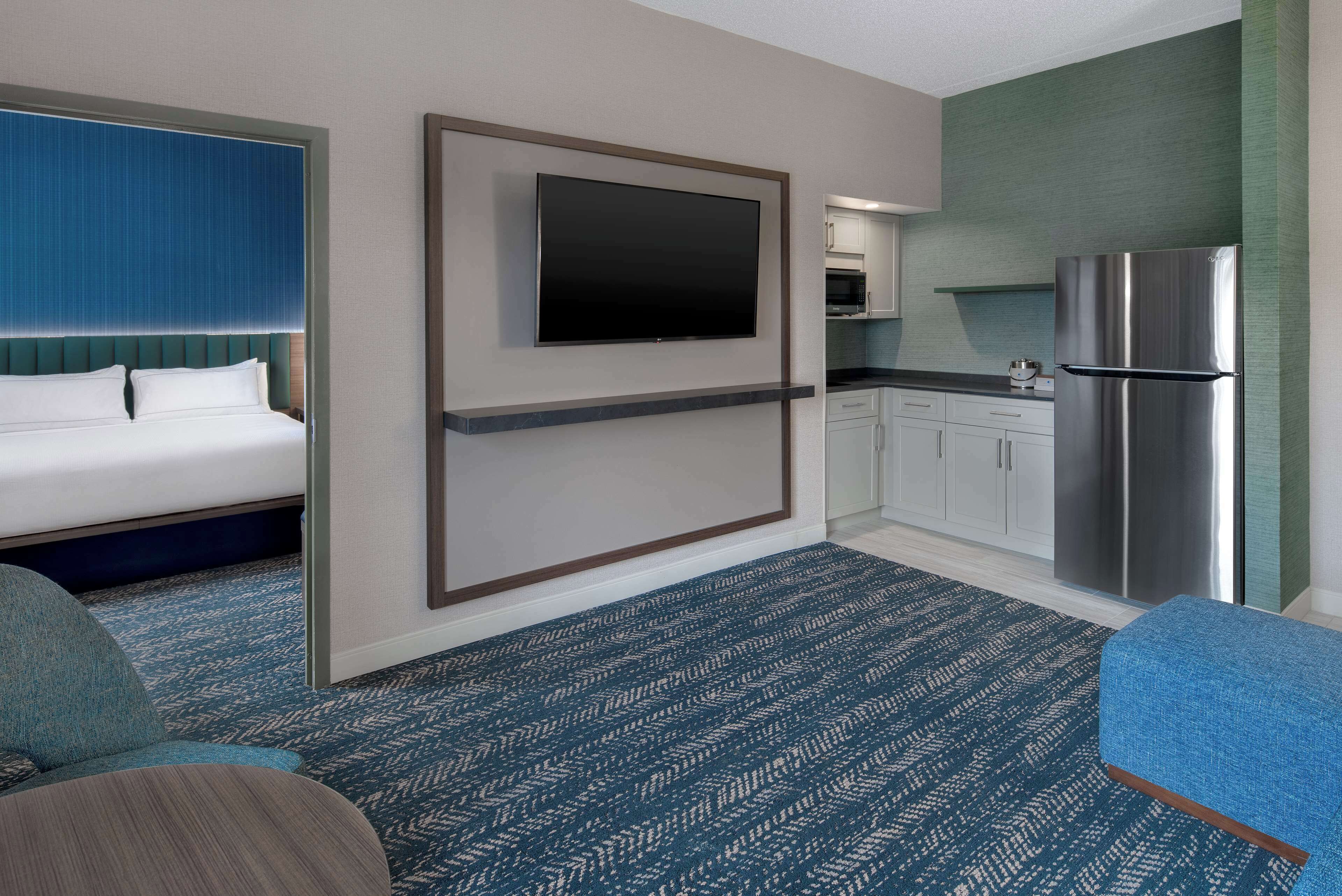 Habitación de hotel con cama, televisor montado en la pared, kitchenette con refrigerador y alfombra estampada.