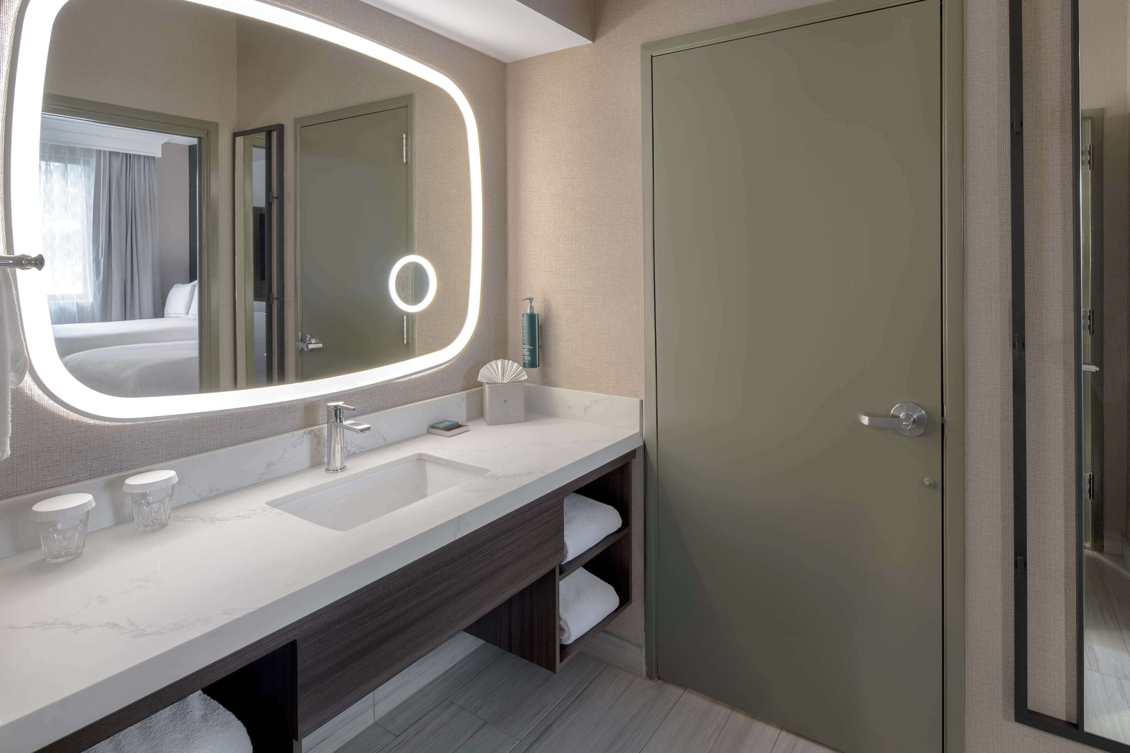 A modern bathroom with a large mirror, a sink, and a toilet. There is a towel rack and a bottle of hand soap on the counter.