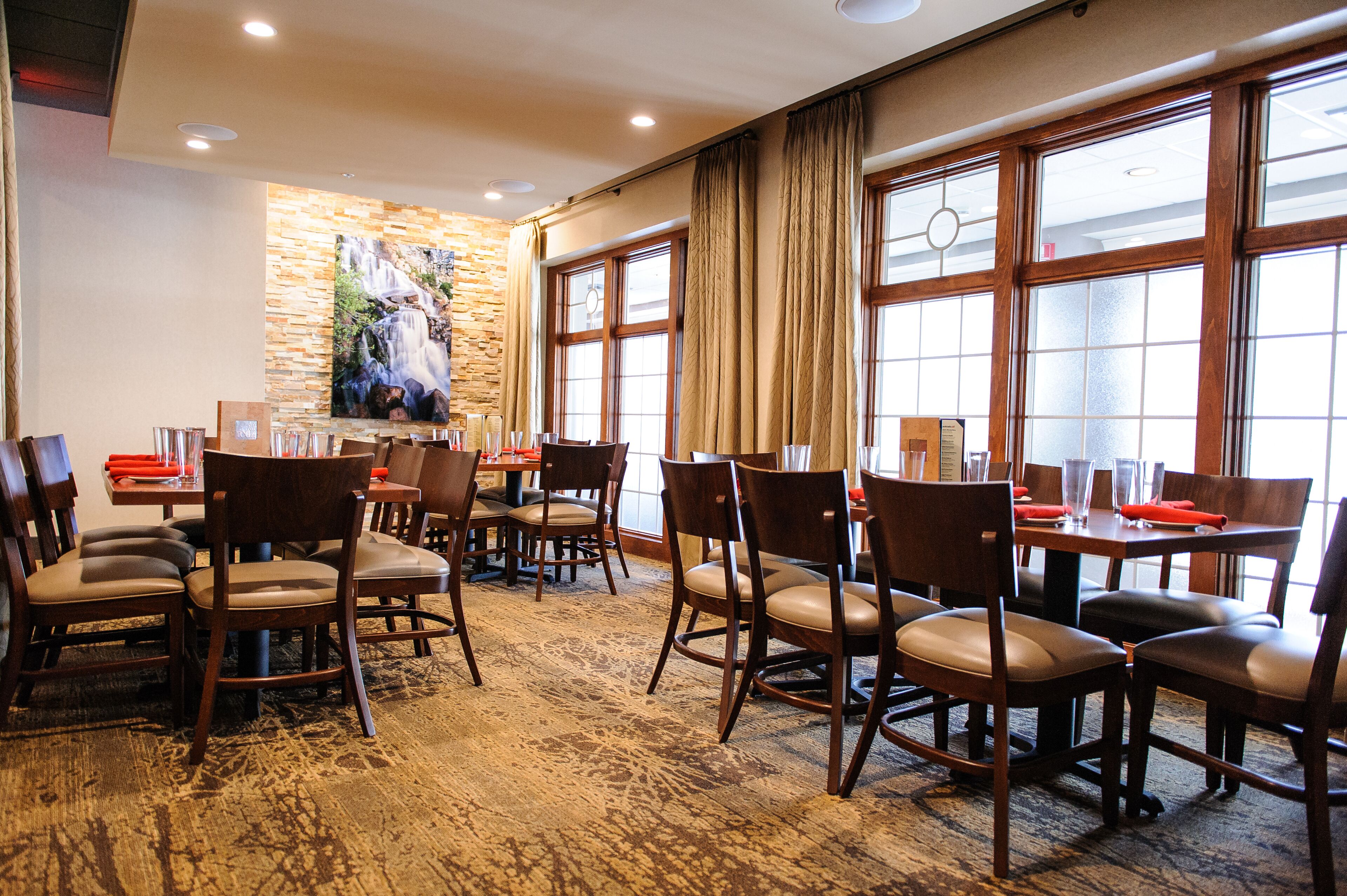 A dining area with multiple tables and chairs, large windows with curtains, and a stone wall with a painting.