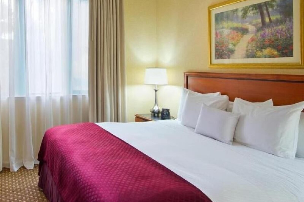 A hotel room with a bed, a nightstand, a lamp, a painting on the wall, and a window with curtains.