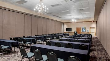 A conference room with rows of tables and chairs, a chandelier, a flat-screen TV, and a painting on the wall.