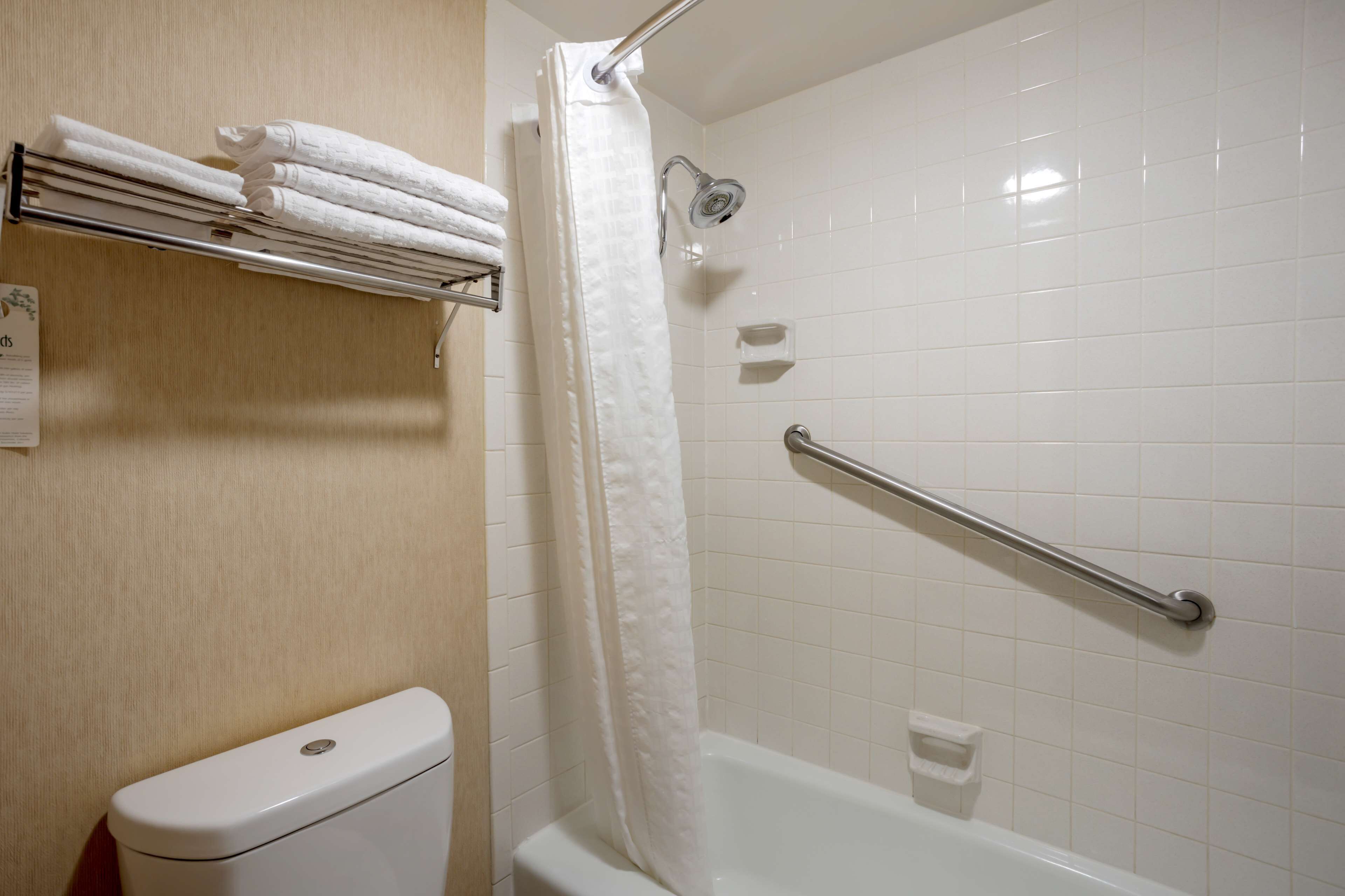 Un baño con inodoro blanco, ducha con cortina blanca, barra de apoyo y toallero con toallas dobladas.
