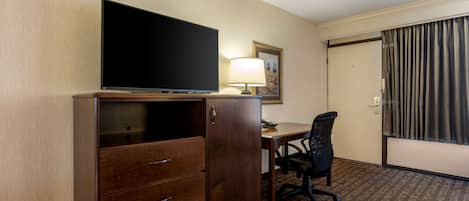 A hotel room with a flat-screen TV on a wooden cabinet, a desk with a chair, a lamp, and a door with curtains.