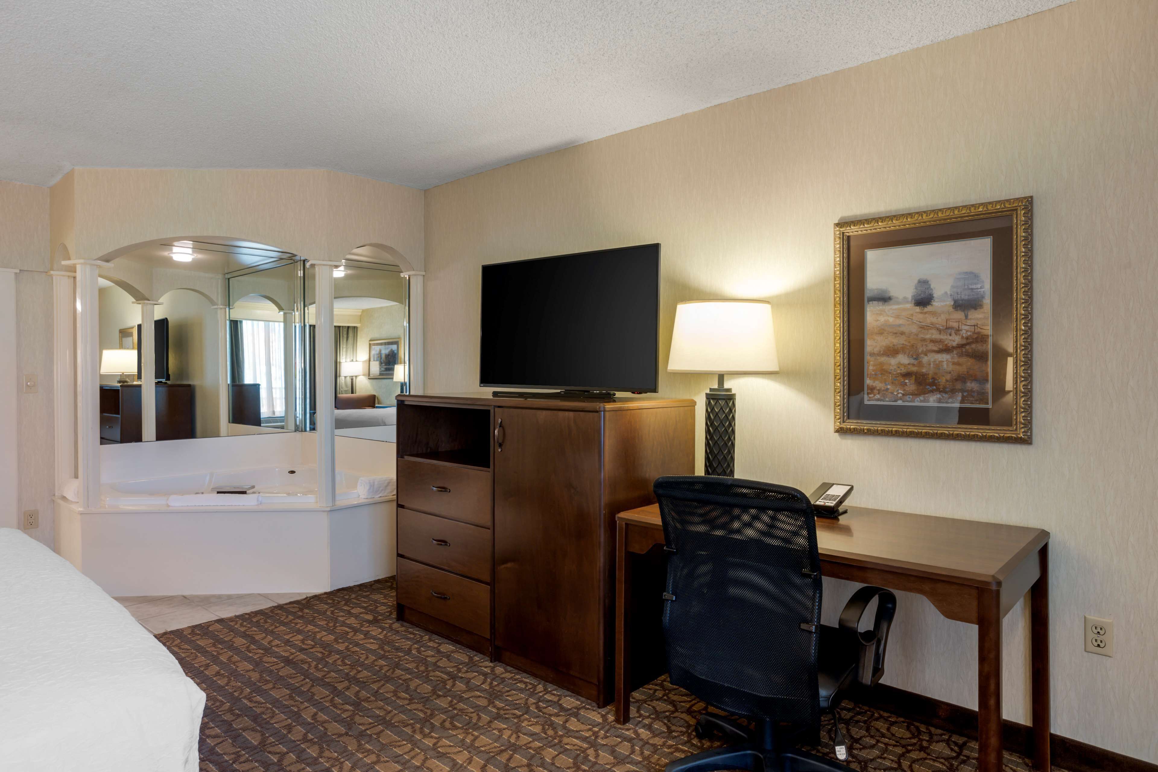 A hotel room with a bed, a desk with a chair, a television, a lamp, a dresser, and a painting on the wall.