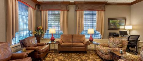 A hotel room with a brown leather sofa, patterned armchairs, a wooden coffee table, and a large window with blinds.