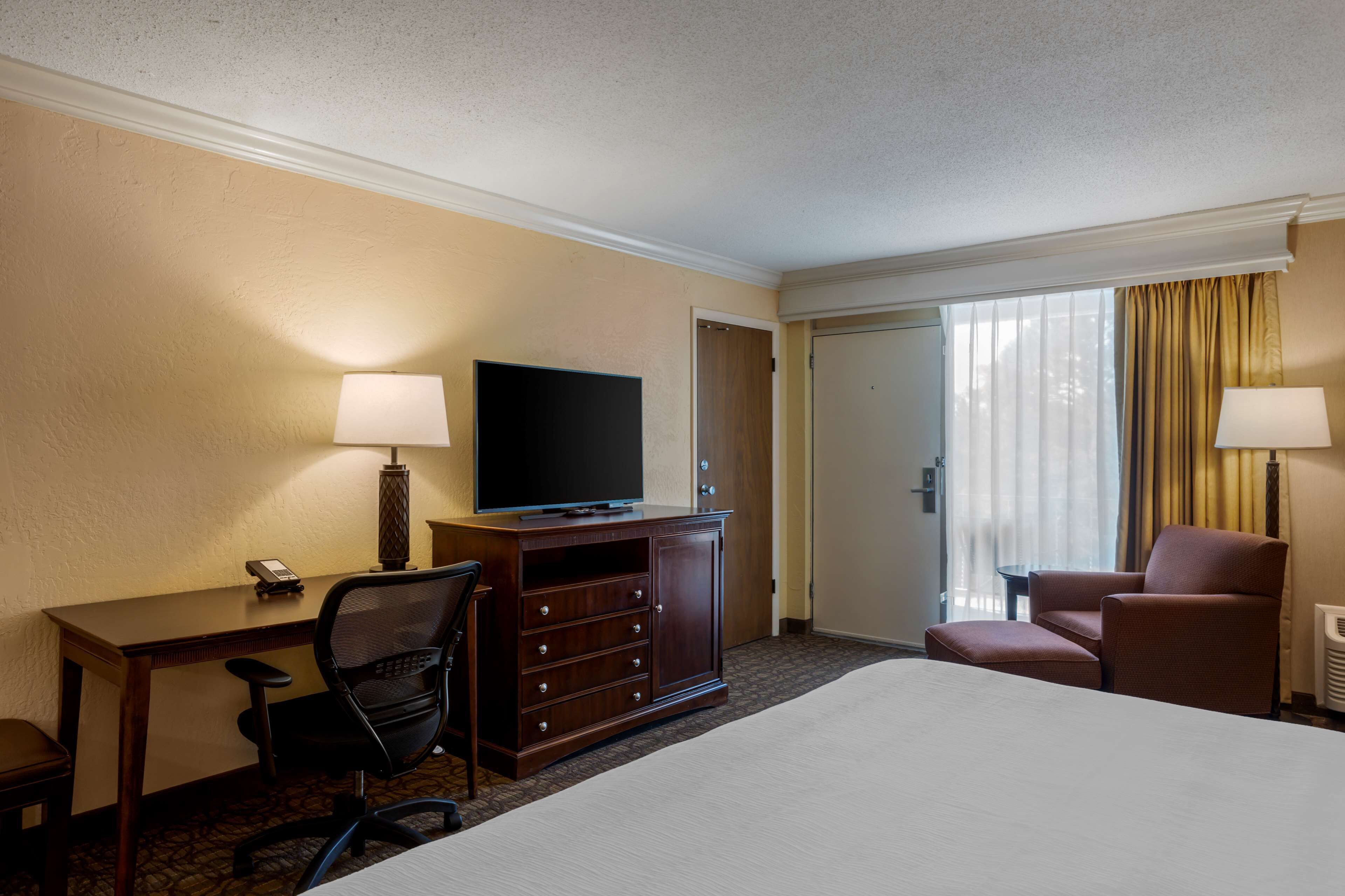 A hotel room with a large bed, a desk with a television, a chair, a dresser, and a window with curtains.
