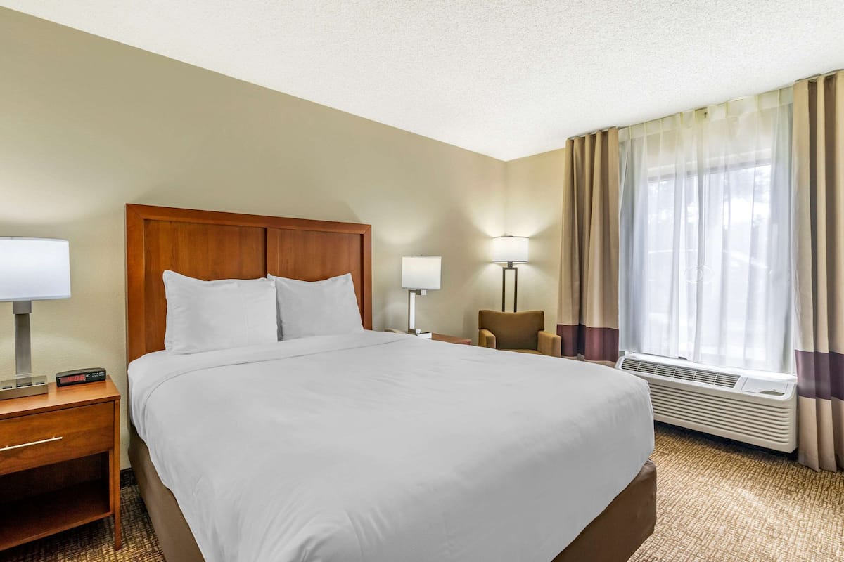A hotel room with a large bed, a nightstand with a lamp, a chair, and a window with curtains.