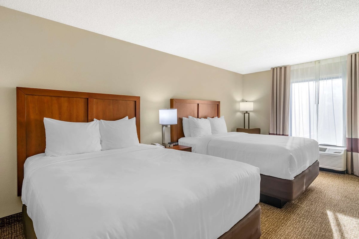 A hotel room with two beds, wooden headboards, white bedding, a bedside table with a lamp, and a large window with curtains.