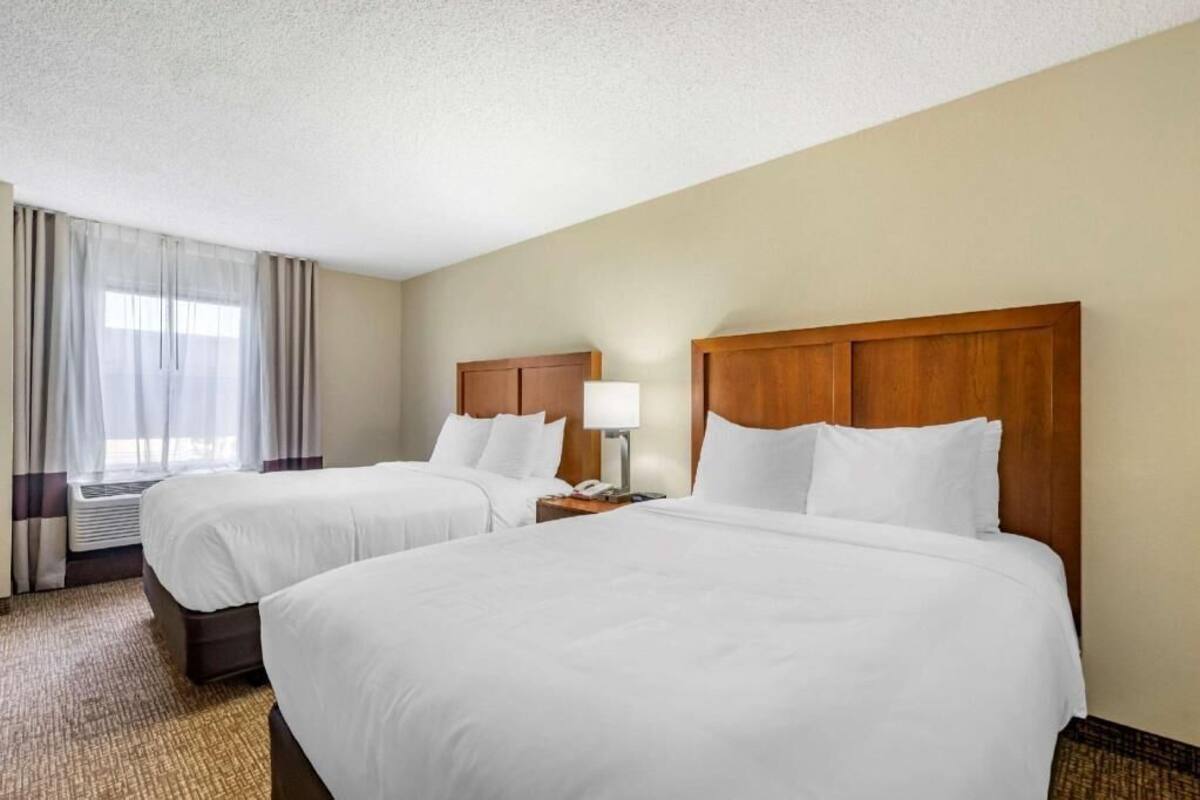 A hotel room with two beds, a wooden headboard, a bedside table with a lamp, and a window with curtains.