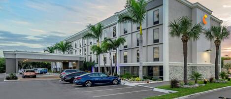 A hotel with a modern design, featuring a parking lot with several cars and palm trees.