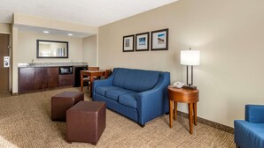A hotel room with a blue sofa, a wooden side table with a lamp, a small ottoman, a kitchenette with a microwave, and a framed picture on the wall. - Comfort Inn & Suites St. Pete - Clearwater International Airport (Clearwater)