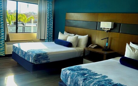 A hotel room with two beds, a large window with curtains, a wooden headboard, and a bedside table with a lamp.