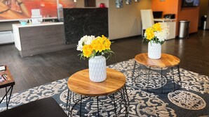 Un hall d’hôtel avec un comptoir d’accueil, deux tables rondes en bois ornées de vases de fleurs, une moquette à motifs et des œuvres d’art accrochées aux murs.