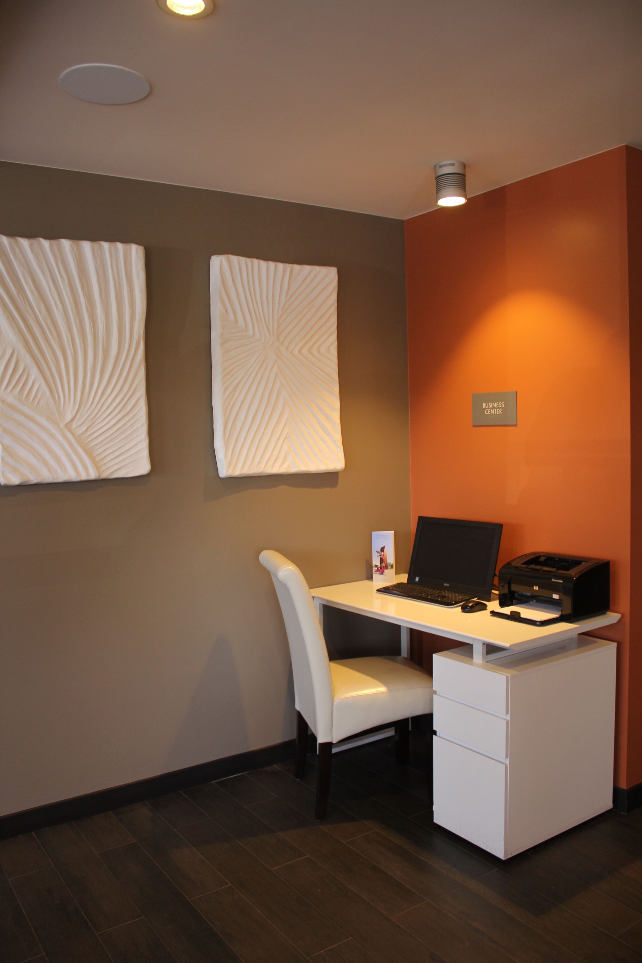A modern office space with a white desk, a laptop, a printer, and a chair. There are two wall-mounted light fixtures and a sign that reads "BUSINESS CENTER".
