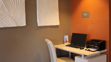 A modern office space with a white desk, a laptop, a printer, and a chair. There are two wall-mounted light fixtures and a sign that reads "BUSINESS CENTER".
